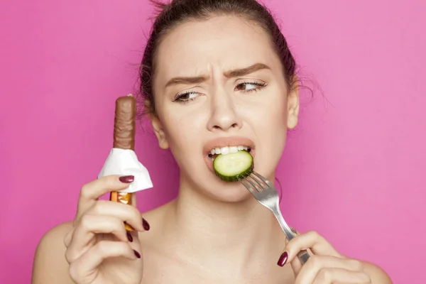 年轻漂亮的女人吃黄瓜和看在粉红色的背景巧克力 — 图库照片