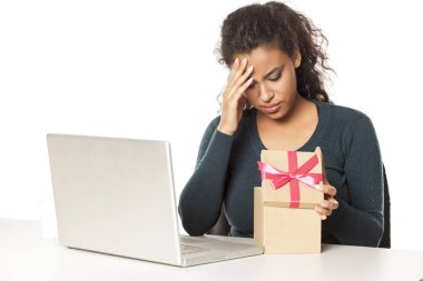 unhappy african woman at the desk with a laptop opens a box with a gift clipart