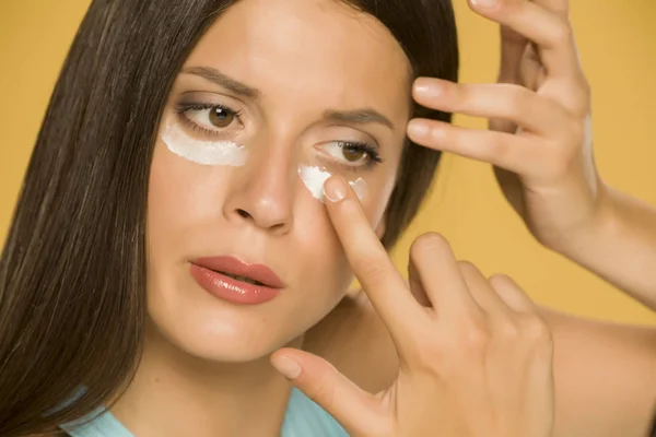 Young Woman Applying Creme Her Low Eyelids Yellow Background — Stock Photo, Image