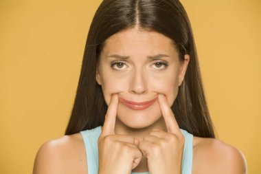 Young sad woman forcing her smile with her fingers on yellow background clipart