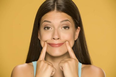 Young woman forcing her smile with her fingers on yellow background clipart