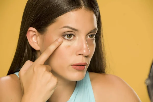 Young Woman Applying Concealer Her Low Eyelids Yellow Background — Stock Photo, Image