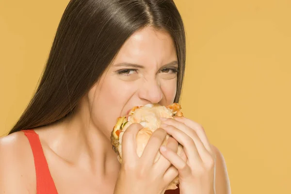 黄色の背景にハンバーガーを食べる若い貪欲な女性 — ストック写真