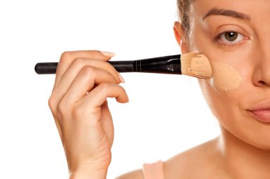 Beyaz arka planda fırçayla sıvı temel uygulayan genç bir kadın.