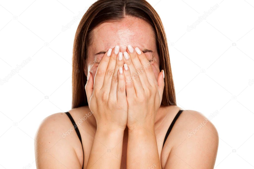 A young happy and shy woman covers her face with her hands on white background