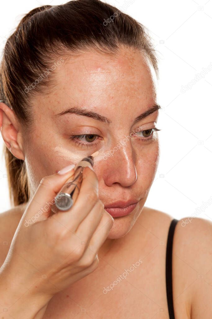 Young woman applying concealer under her eyes with brush on white background