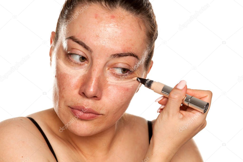 Young woman applying concealer under her eyes with brush on white background