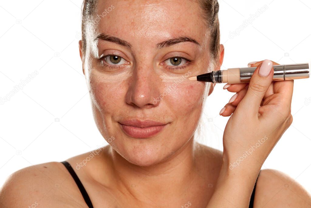 Young woman applying concealer under her eyes with brush on white background