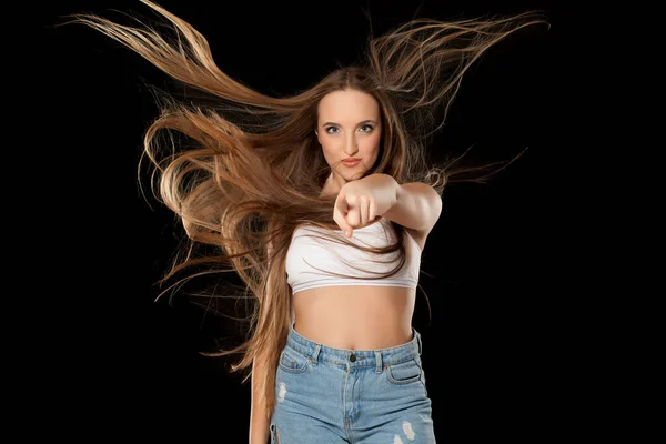 Young woman with very long flying hair on black background pointing on you