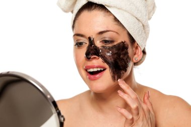 Young beautiful woman with towel on her head applying black mask on her face clipart