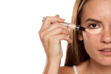 Genç ve güzel bir kız, beyaz arka planda gözlerinin altına kapatıcı sürüyor.