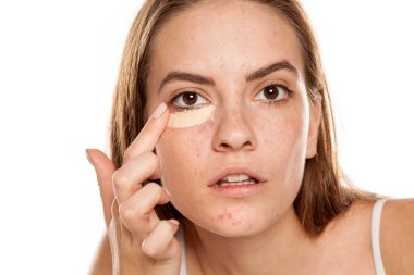 Genç ve güzel bir kız, beyaz arka planda parmakları gözlerinin altında kapatıcı sürüyor.