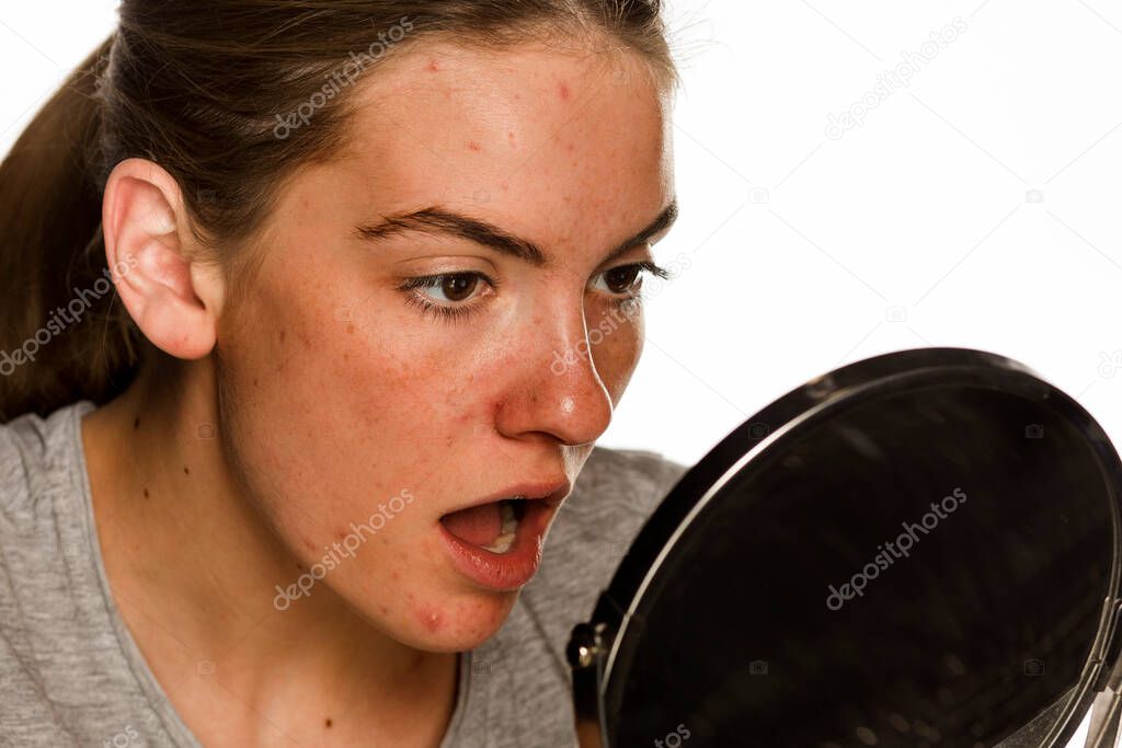 Young concerned woman without makeup looking her self on the mirror on white background