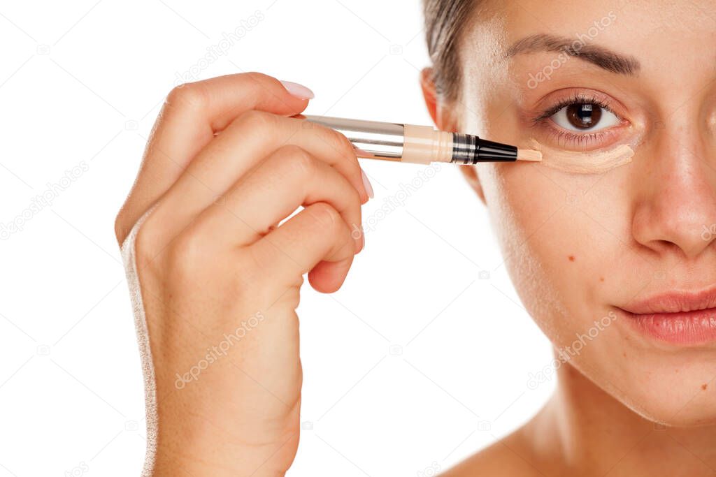 young beautiful girl applying concealer under her eyes on white background