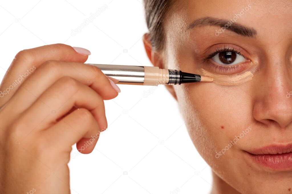 young beautiful girl applying concealer under her eyes on white background