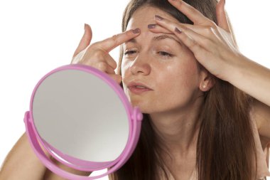 Makyajsız genç bir kadın aynaya bakar ve sivilcelerini sıkar.