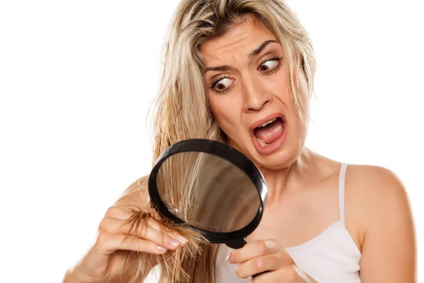 头发凌乱的金发女人震惊极了 她用白色背景的放大镜看她的分叉头 — 图库照片