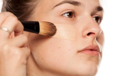 Beyaz arka planda fırçayla sıvı temel uygulayan genç bir kadın.