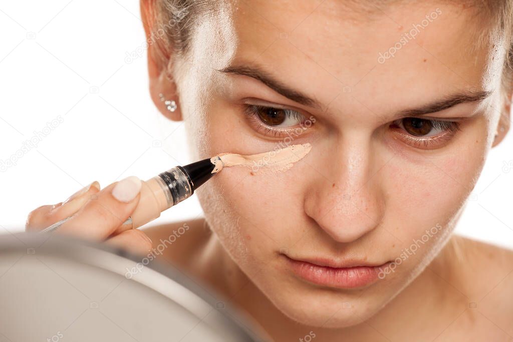 Young woman applying concealer under her eyes on white background