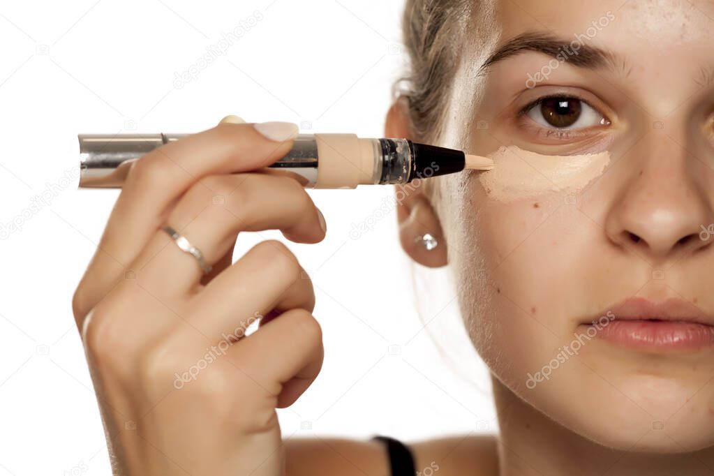 Young woman applying concealer under her eyes on white background