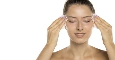 Makyajsız, makyajsız genç bir kadının beyaz arka planda pamuk pedlerle makyajı temizlerken çekilmiş bir fotoğrafı.