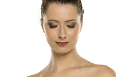 Kahverengi saçlı, makyajlı, gözleri kapalı ve aşağı bakan beyaz arka planda genç bir kadının stüdyo fotoğrafı.