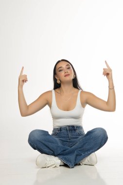 Uzun siyah saçlı, beyaz kolsuz bluzlu, kot pantolonlu ve spor ayakkabılı, çapraz bacaklı ve yukarı doğru beyaz arkaplana bakarken iki elini havaya kaldırmış genç bir kadının stüdyo fotoğrafı.