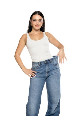 Stüdyo resmi, uzun siyah saçlı, beyaz kolsuz bluzlu, bol kot pantolonlu, beyaz arka plana uyacak şekilde bel bandı tutan genç bir kadın.