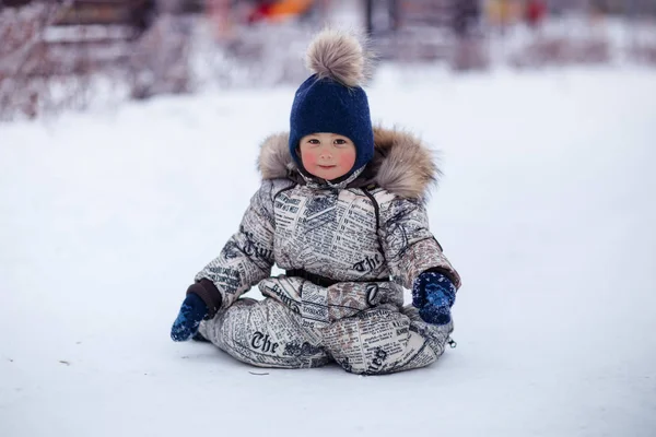 Wesoły Mały Chłopiec Siedzi Pokrytej Śniegiem Ziemi Podwórku Patrząc Kamery — Zdjęcie stockowe