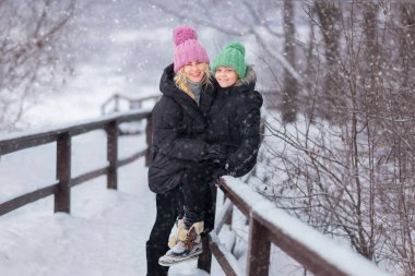 mutlu anne karlı parkta ahşap köprü korkuluk oturan oğlu ile kameraya bakıyor 
