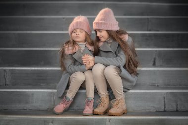 merdivenlerde oturan ve el ele tutuşarak pembe örgü şapka giymiş neşeli kız kardeş 