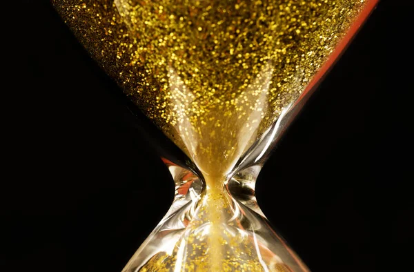 Sand and golden glitter passing through the glass bulbs of an hourglass measuring the passing time as it counts down to a deadline or closure on a black background with copy space
