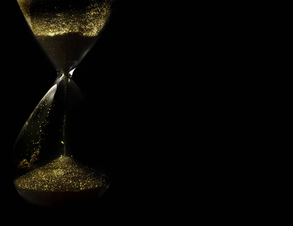 Sand and golden glitter passing through the glass bulbs of an hourglass measuring the passing time as it counts down to a deadline or closure on a black background with copy space