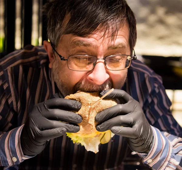 Uomo anziano mangiare gustosi guanti neri hamburgerin — Foto Stock