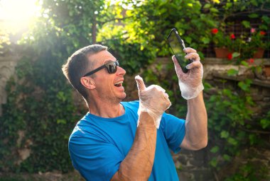 Tek kullanımlık eldivenli gülümseyen adam dışarıda akıllı telefon tutuyor.