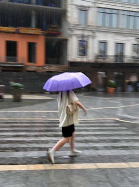 Şiddetli yağmurda caddede hızla yürüyen bir kadın, elinde hareket halinde büyük bir şemsiye, bulanık.