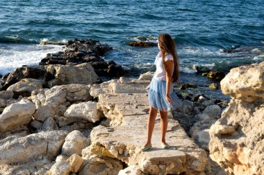 Genç, güzel, beyaz, açık deniz kıyısında, kumsalda, büyük sarı taşların arasında tatlı bir kız.