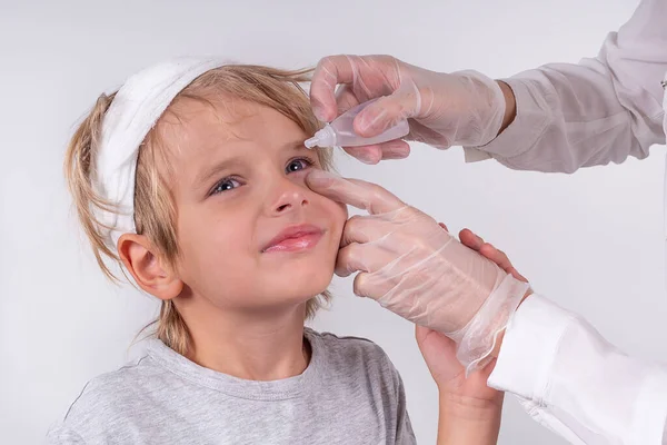 Female hands optometrist wearing transparent medical gloves putting eye drop patient little blonde boy eyes in ophthalmology clinic. Sick look. White background. — Stock Photo, Image