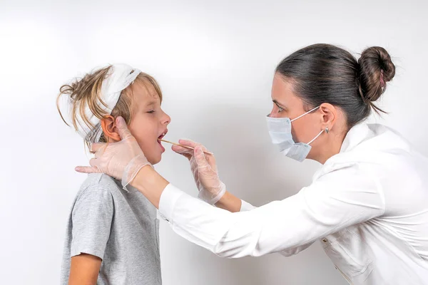 Medicine, healthcare and pediatry concept. A female doctor in medical mask and transparent gloves examines a sick little boy with head injury. Checks the throat and takes a swab for the coronavirus. — Stock Photo, Image