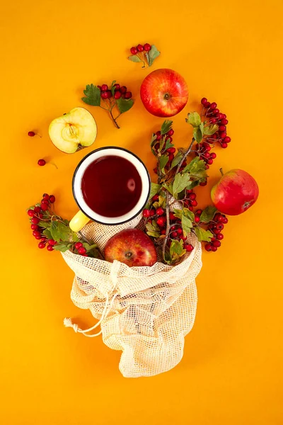 Pommes biologiques de jardin rouge, baies d'aubépine dans un sac de maille et tasse de thé épicé chaud sur un fond orange. Automne nature morte couché plat — Photo