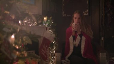 Güzel kız Noel ağacın yanında oturan ve sıcak çikolata içme rahat bir battaniyeye sarılmış