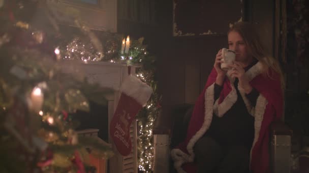 美丽的女孩包裹在一个舒适的毯子坐在圣诞树旁边 喝热巧克力 — 图库视频影像
