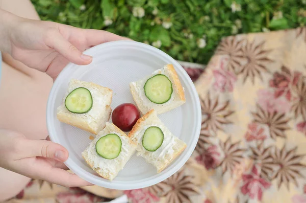A young girl holds a plate with four small sandwiches with melted cheese ,cucumber slices and one plum in the center..Healthy summer picnic in the Park or in the garden. — Stock Photo, Image