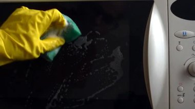Koruyucu eldivenli bir adam mikrodalga fırının kapalı kapısını solüsyonlu süngerle yıkıyor.