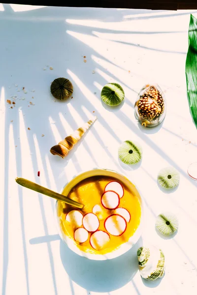 大根のスライスを持つココナッツカボチャのスープ 熱帯植物と白いボードの上に白いボウルに 緑のウニの貝殻とハードシャドウ — ストック写真