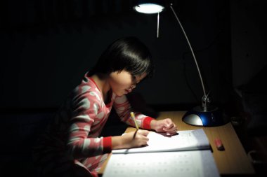 Portrait of little Asian girls doing her homework under the lighting lamp in a dark room clipart