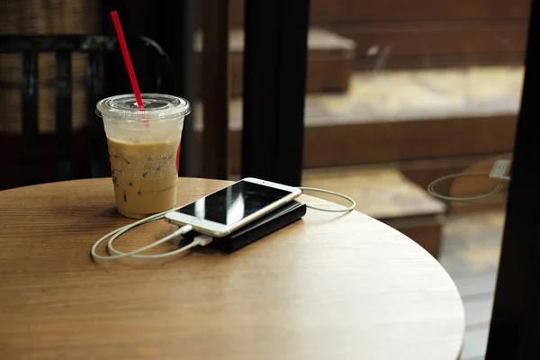 Vintage abstract picture of mobile phone and power bank put on the wooden table with a cup of ice coffee in the coffee shop.