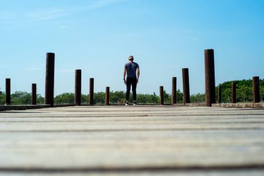 Sıradan giyinmiş yakışıklı bir adamın arka portresi mavi gökyüzü arkaplanlı ahşap bir köprüde duruyor.