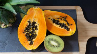 Papaya halves, kiwi, stand stone board with wooden frame, near papai stands the cut off pineapple top, black background, top view clipart