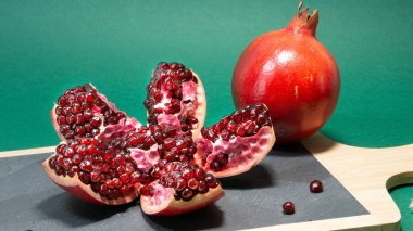 Tahıllar görünür olduğu açık bir nar meyve, başka bir bütün nar meyve yanında duruyor. Ahşap saplı bir taş mutfak tahtası üzerinde meyve standı, arka plan yeşil